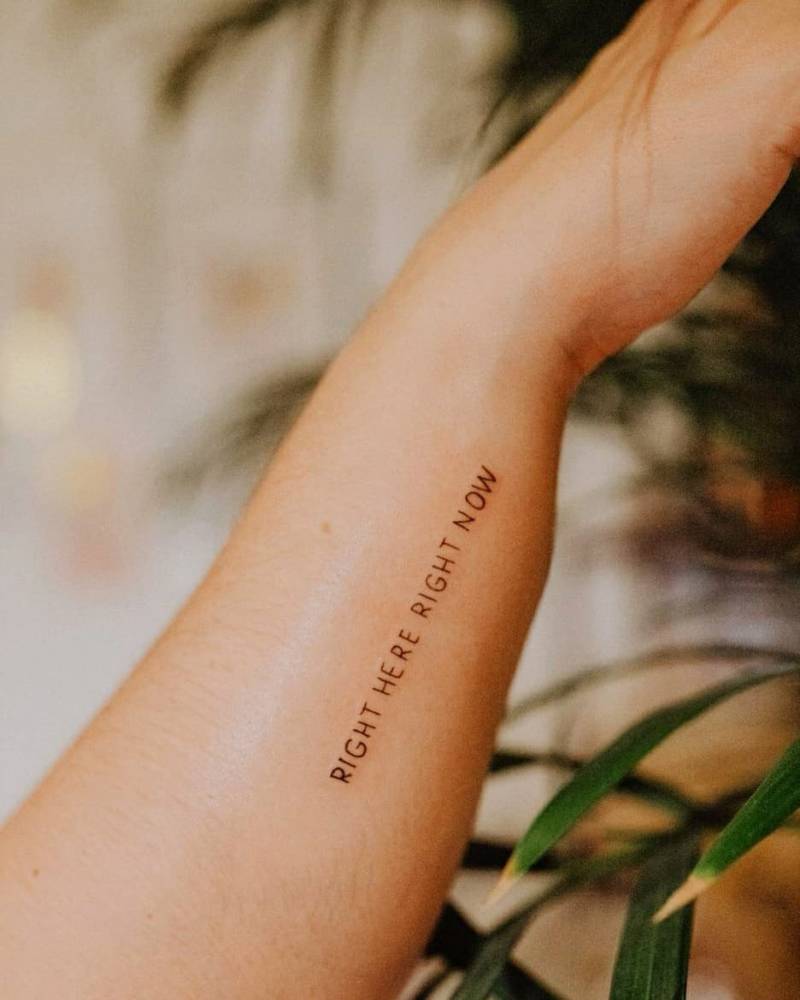 "Right here, right now" lettering tattoo on the forearm.