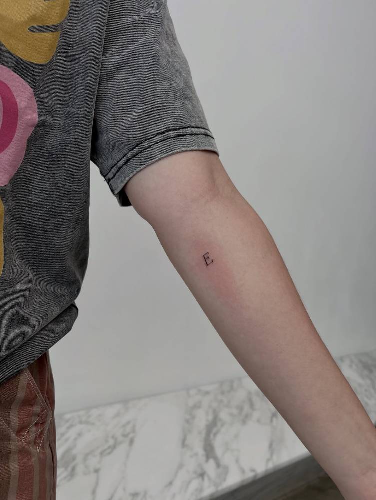 Tiny serif font letter "E" tattoo located on the inner forearm.
