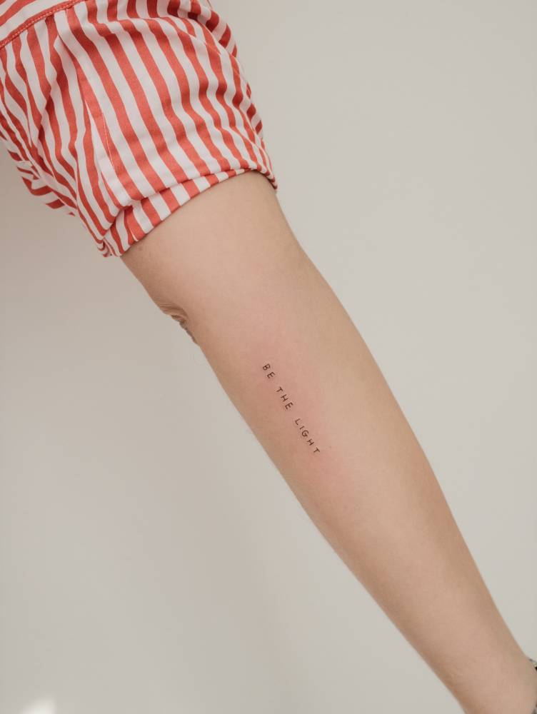 Lettering tattoo that says "be the light" located on the inner forearm.