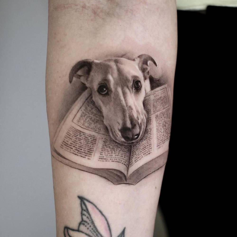Micro-realistic dog and book tattoo on the inner forearm.