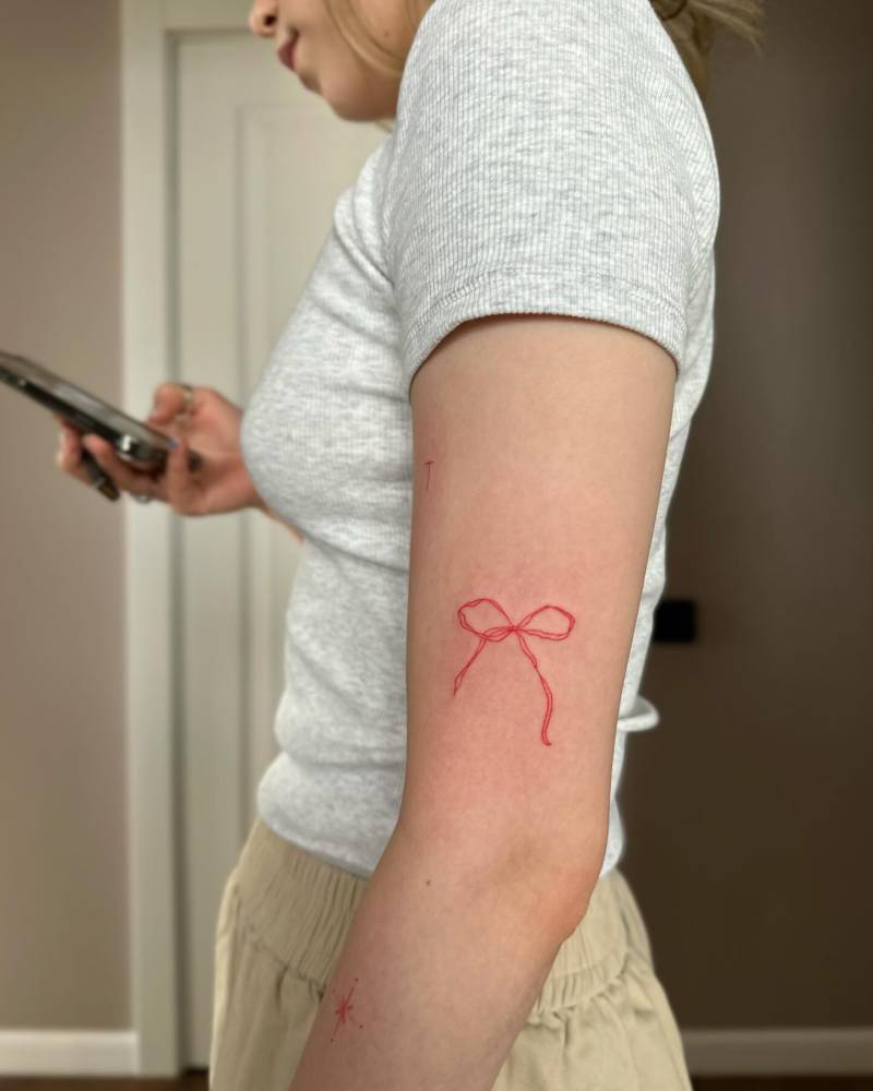 Fine line style red bow tattoo located on the upper arm.