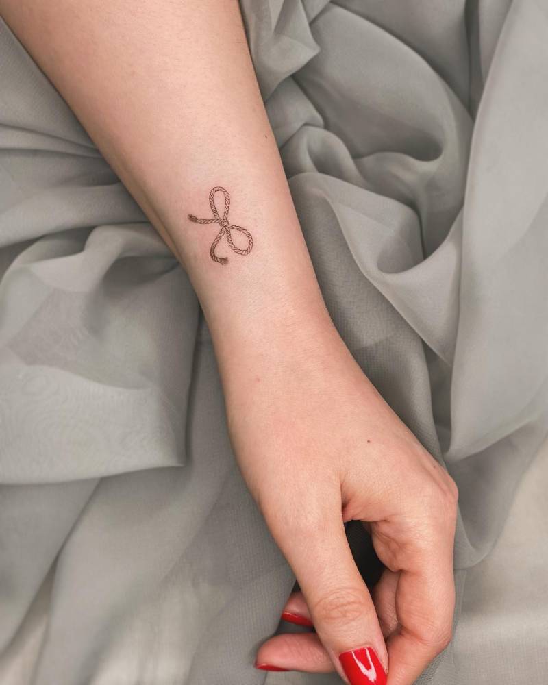 Fine line style rope bow tattoo located on the wrist.
