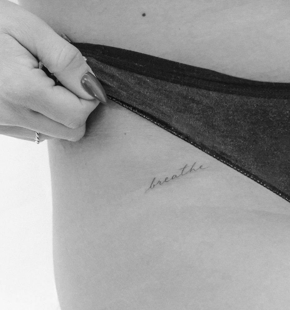 Handwritten lettering tattoo of the word "breathe" placed on the pelvis.