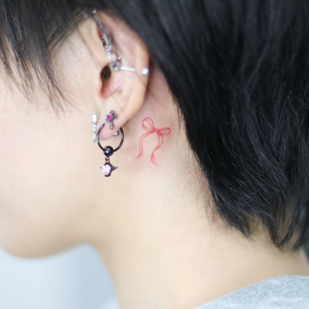 Red bow tattoo in fine line style placed behind the ear.