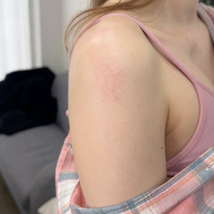 White ink bow tattoo located on the shoulder, minimalistic style.