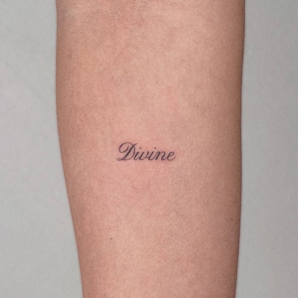 Word "Divine" lettering tattoo located on the inner forearm.