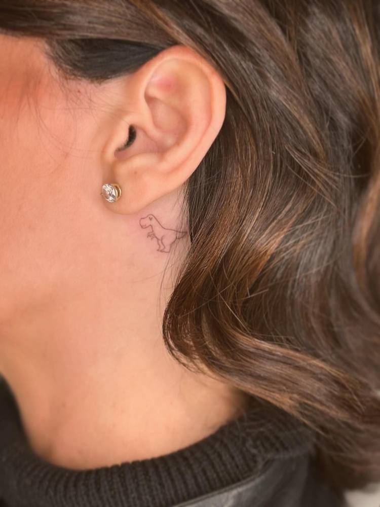 Cute fine line T-Rex tattoo located behind the ear.