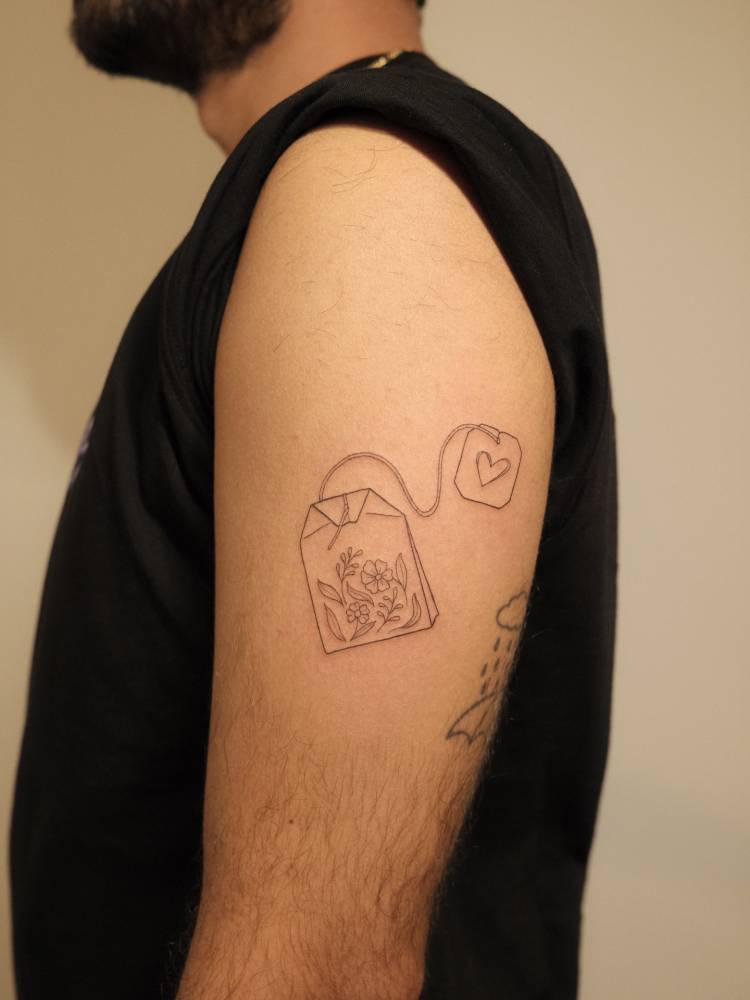 Tea bag tattoo placed on the upper arm, fine line style.