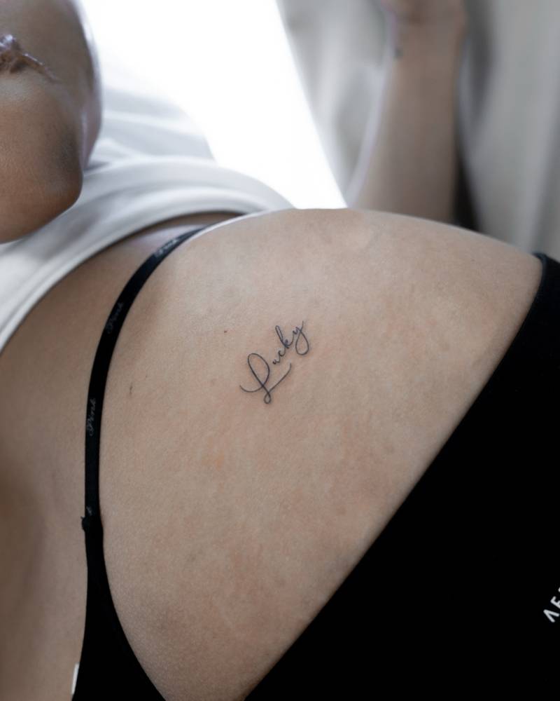 Handwritten font word "lucky" tattooed on the shoulder.