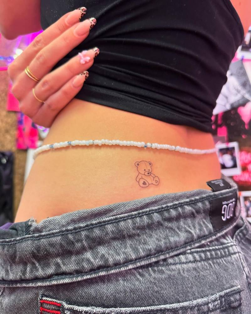 Fine line style teddy bear tattoo located on the lower back.