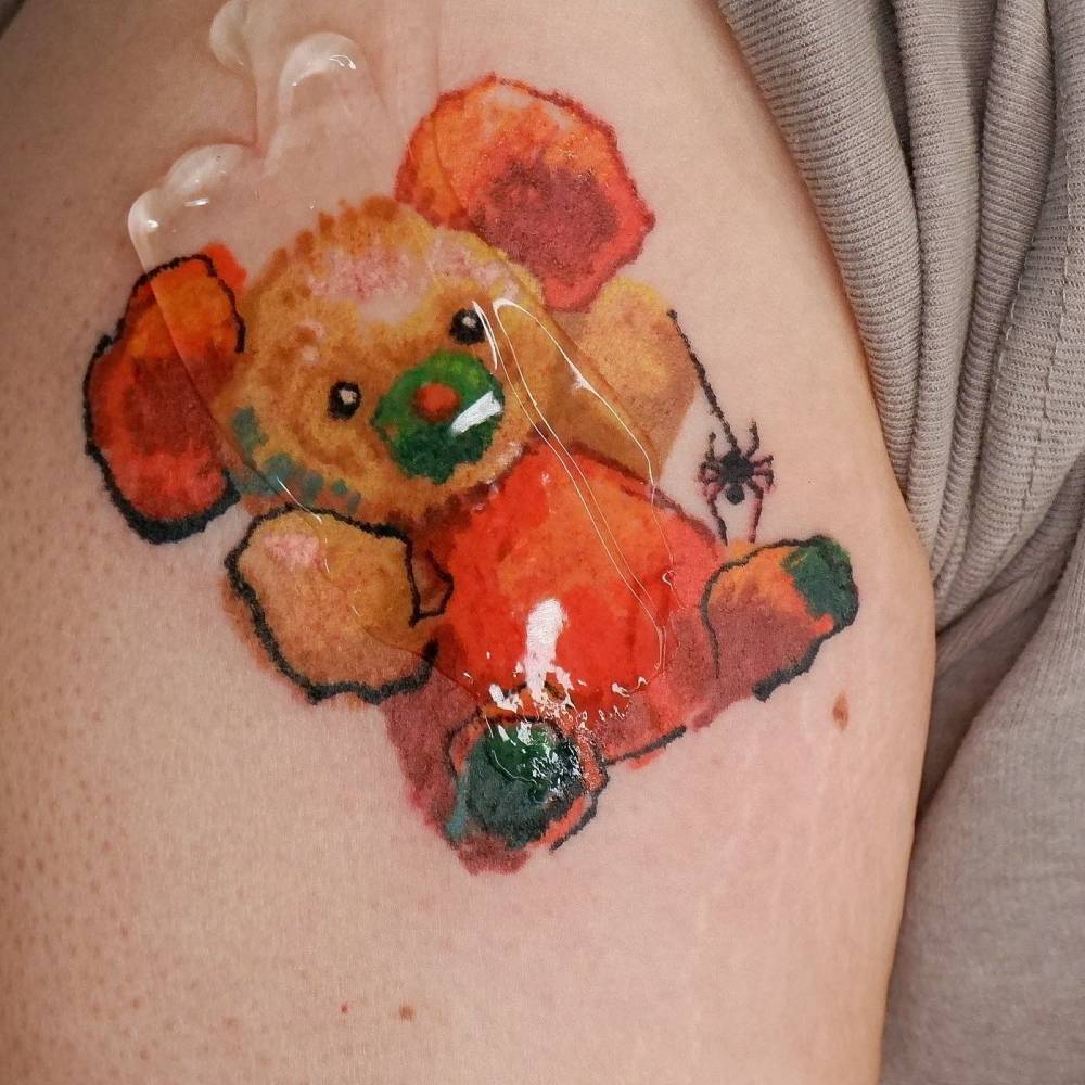Watercolor style teddy bear tattoo located on the upper arm.