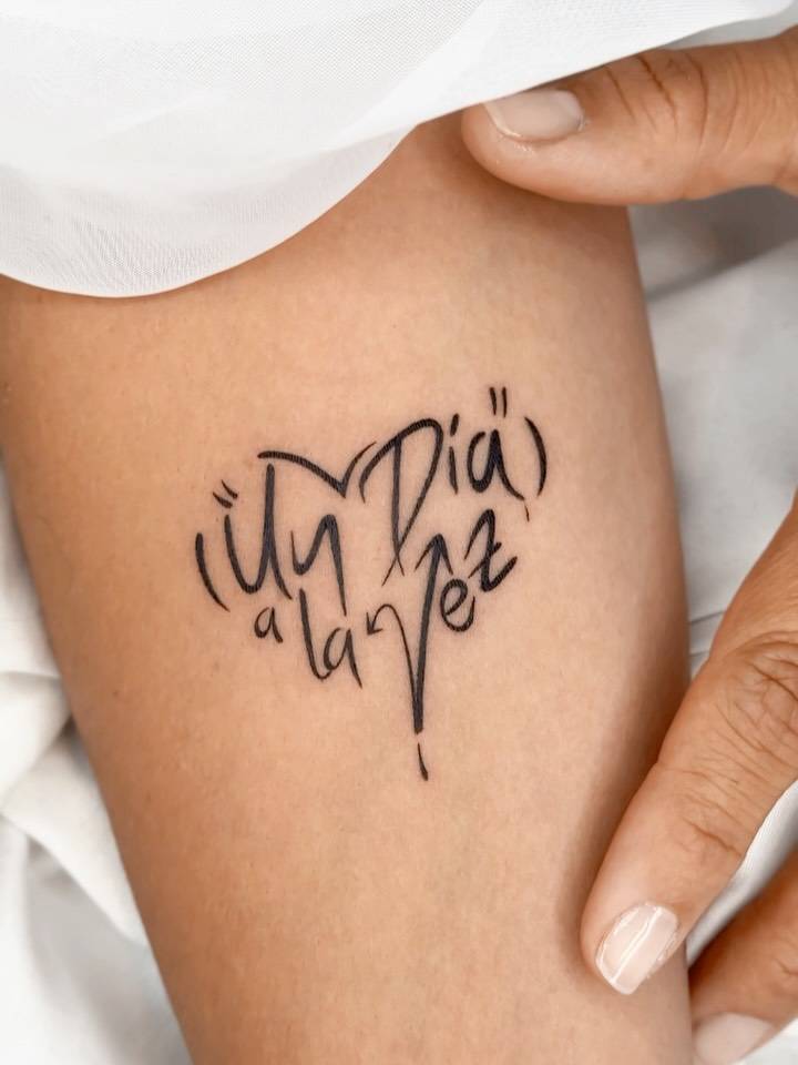 "Un día a la vez" lettering tattoo in a heart shape located on the inner forearm.
