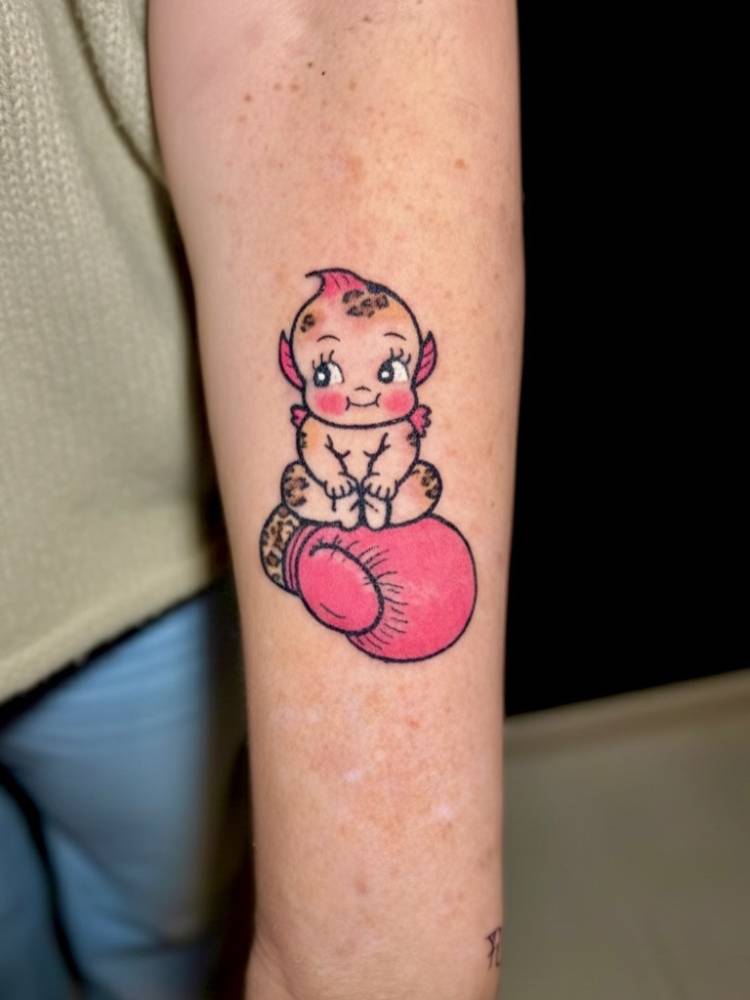 Kewpie and boxing glove tattoo in cartoon style located on the inner forearm.