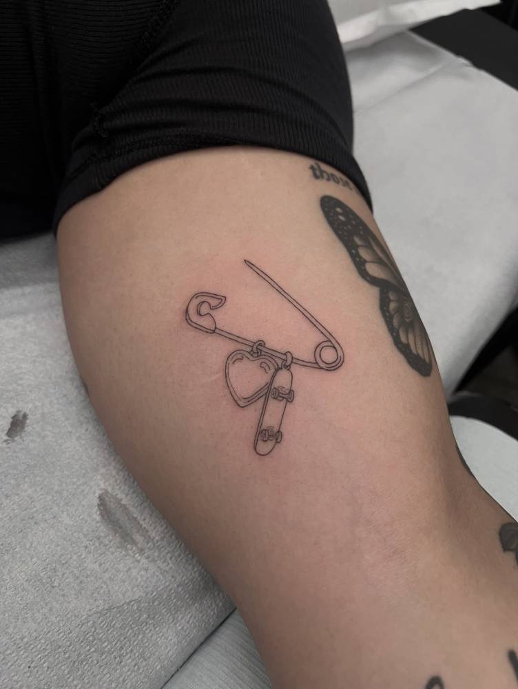 Safety pin and pendants tattooed on the inner arm, fine line style.