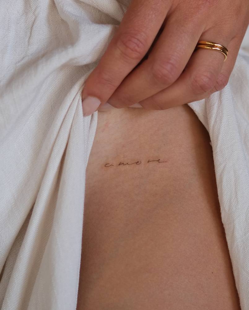 Handwritten lettering tattoo of the word "amore" located on the pelvis.