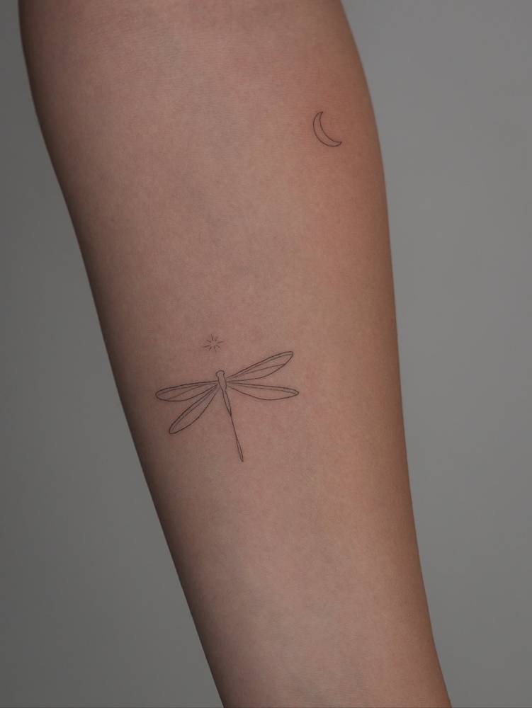 Little dragonfly and crescent moon in fine line style tattooed on the inner forearm.