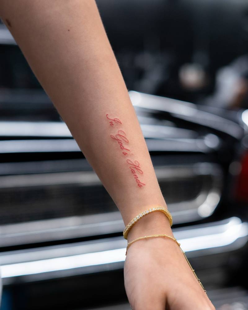 "In God's hands" lettering tattoo in red ink done on the wrist.