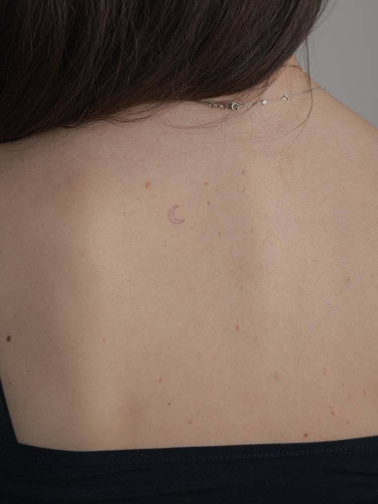 Tiny micro moon tattoo located on the upper back, minimalistic style.