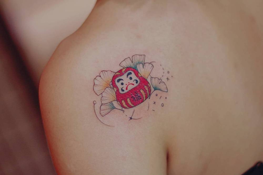 Daruma doll and ginkgo leaves tattoo placed on the shoulder blade.