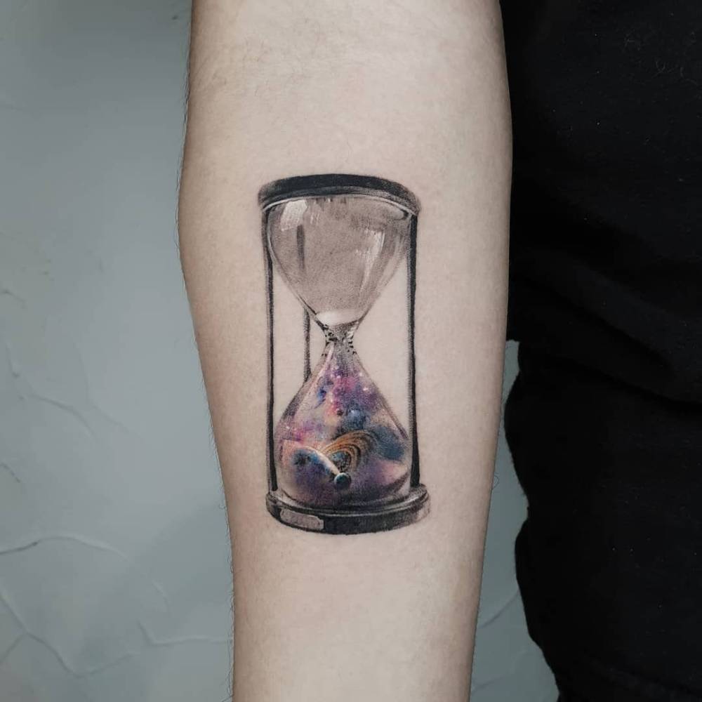 Universe inside the hourglass tattooed on the inner forearm, micro-realistic style.