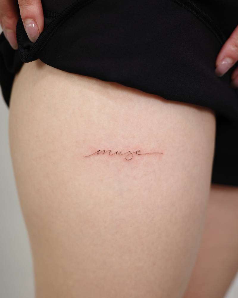Lettering tattoo of the word "muse" handwritten on the thigh.