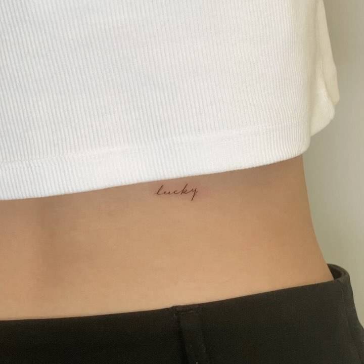 Handwritten word "lucky" lettering tattoo placed on the lower back.