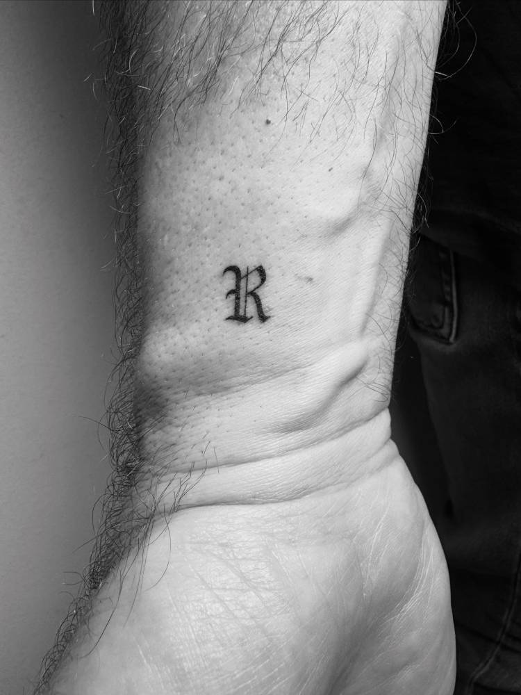 Gothic font letter "R" tattoo placed on the wrist.