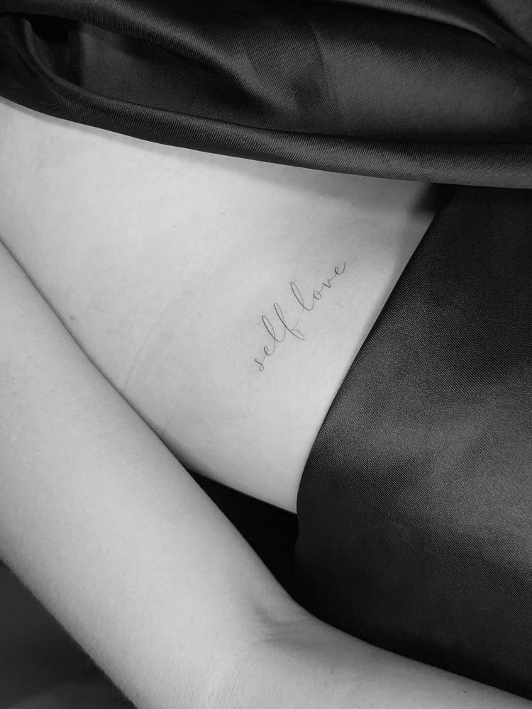 Handwritten font "self love" lettering tattoo located on the rib.