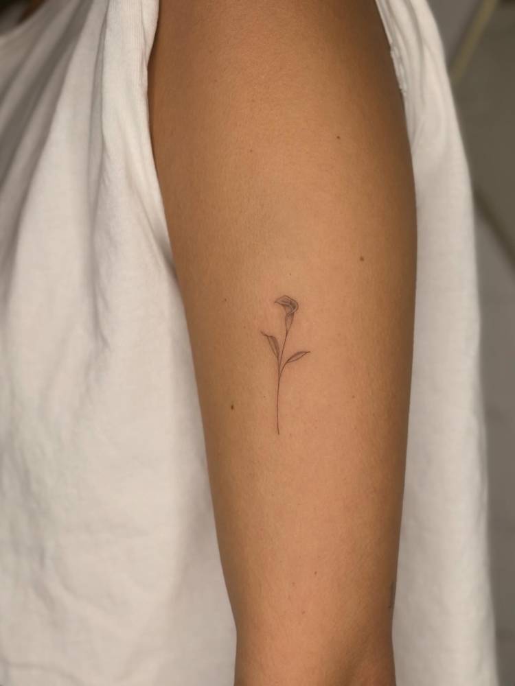 Tiny calla lily flower tattooed on the upper arm, fine line style.