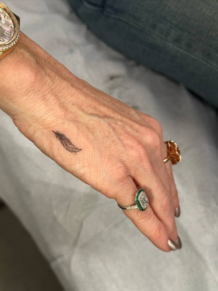 Little feather tattoo in illustrative style located on the hand.