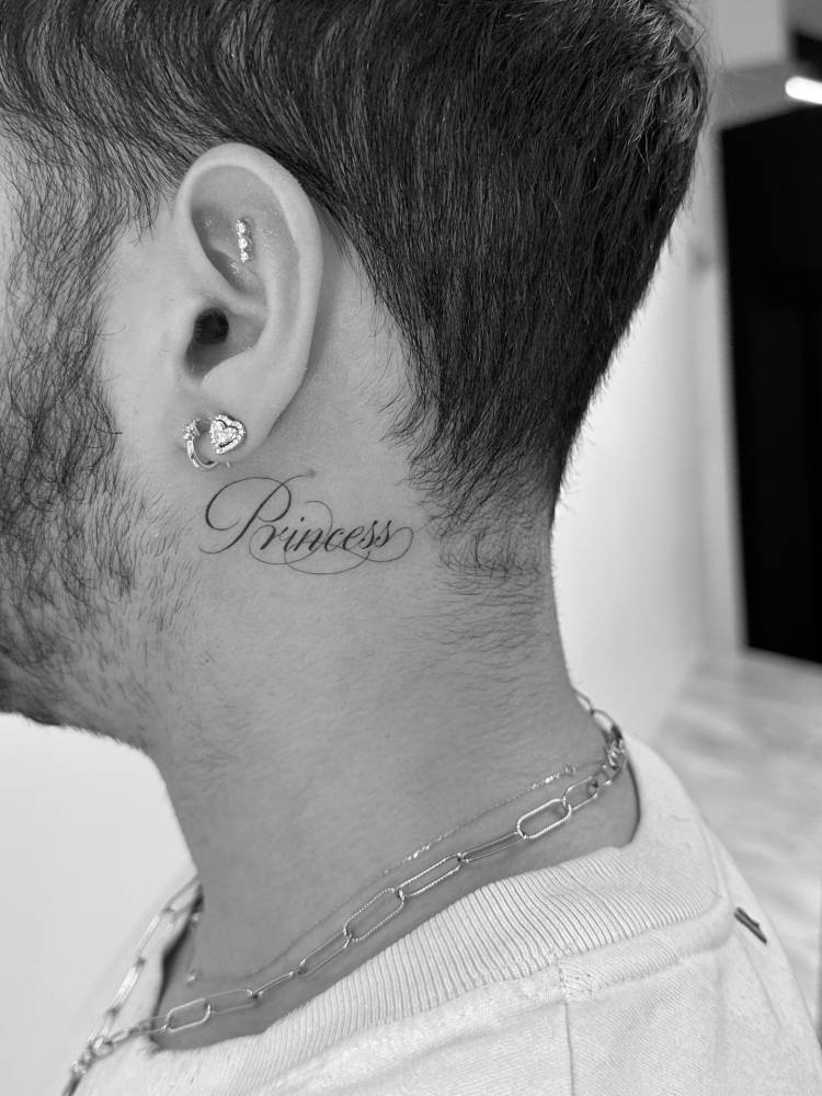 "Princess" lettering tattoo in script font located behind the ear.