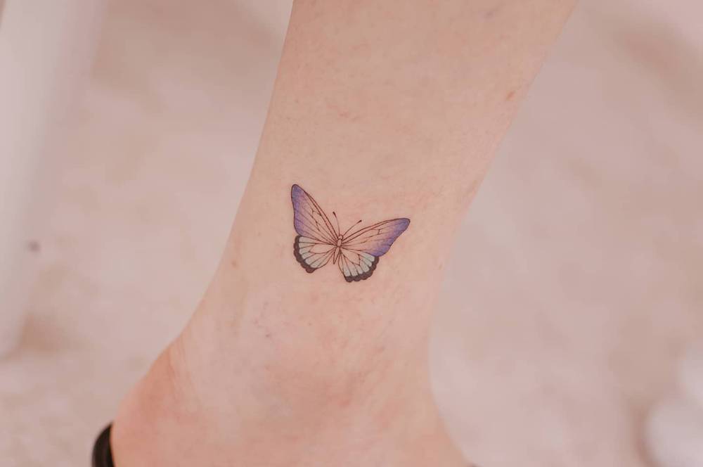 Purple butterfly tattoo in illustrative style located on the ankle.