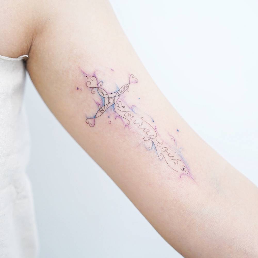 Watercolor "courageous" cross tattoo on the bicep.