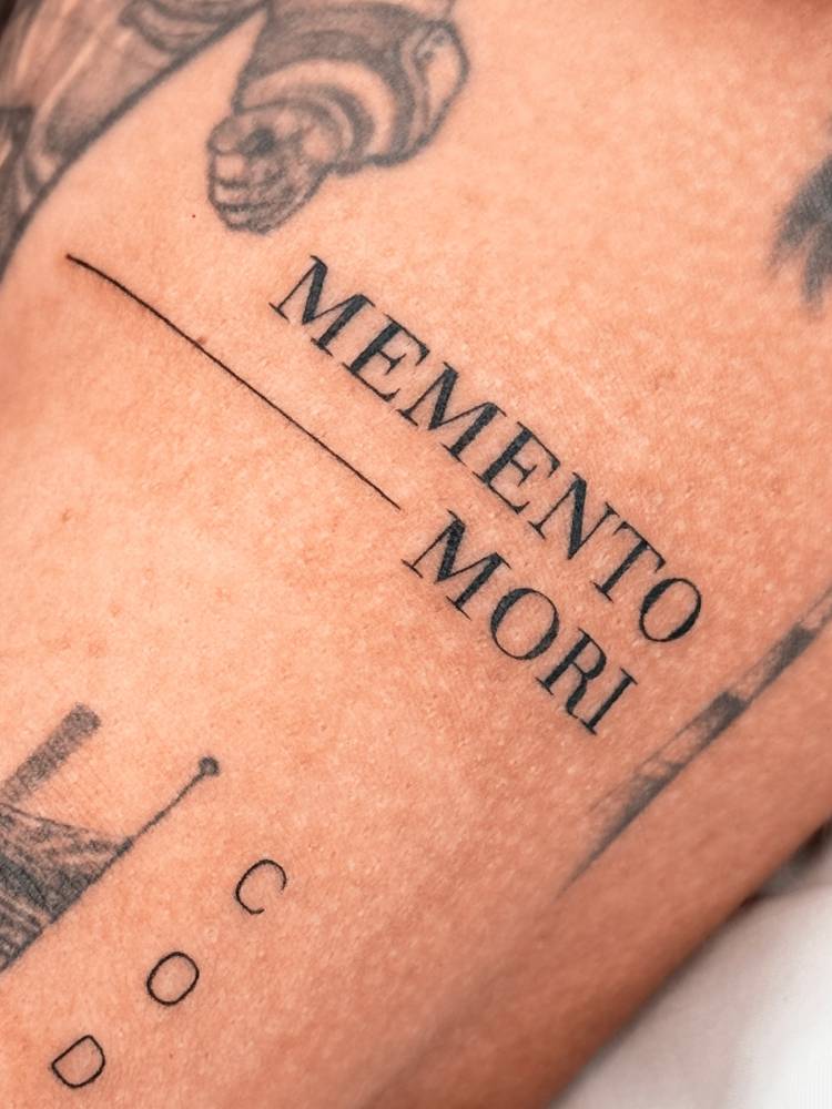 "Memento mori" lettering tattoo in serif font located on the upper arm.