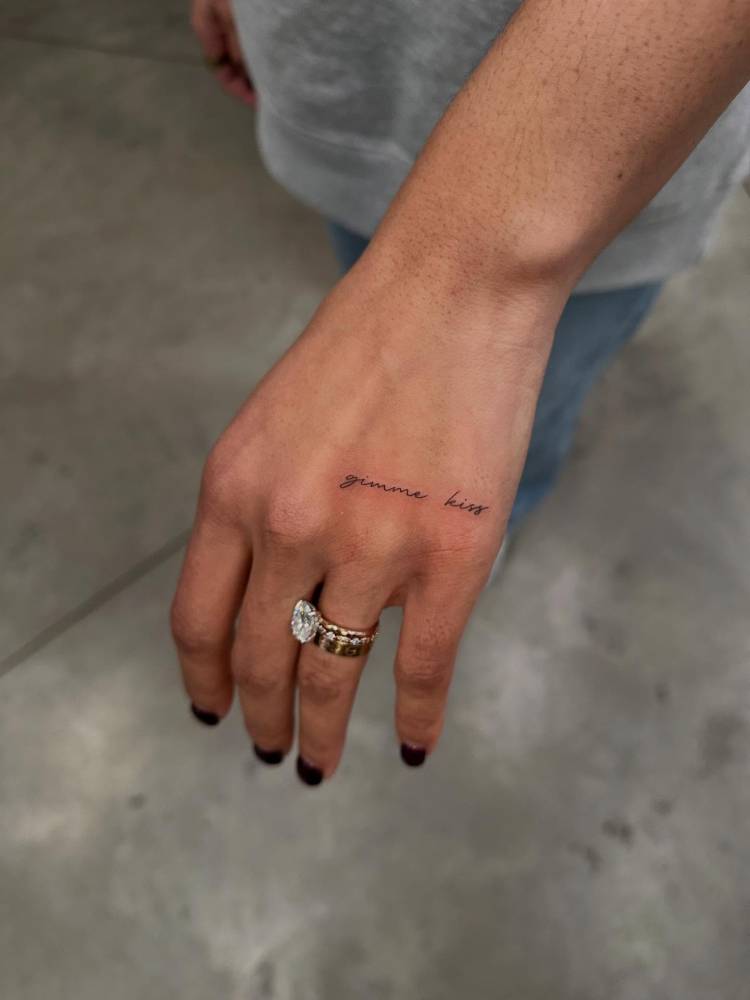 "Gimme kiss" lettering tattoo located on the hand.