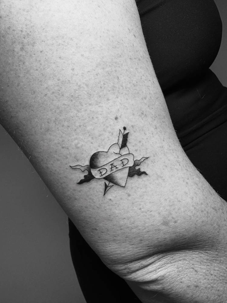 Heart and arrow with banner tattooed on the upper arm.