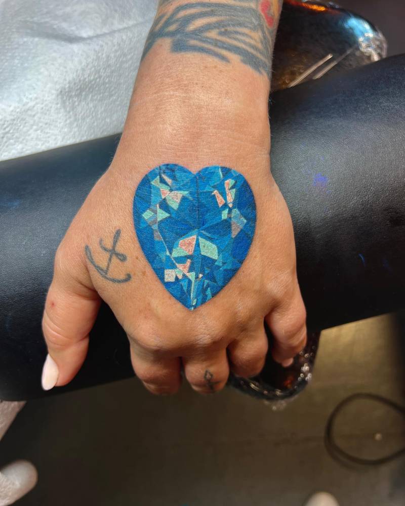 Illustrative style sapphire heart tattoo placed on the hand.