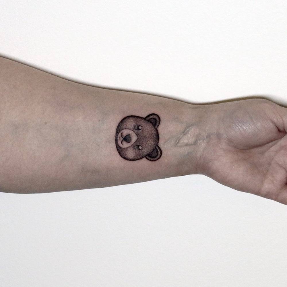 Teddy bear tattoo located on the wrist, illustrative style.