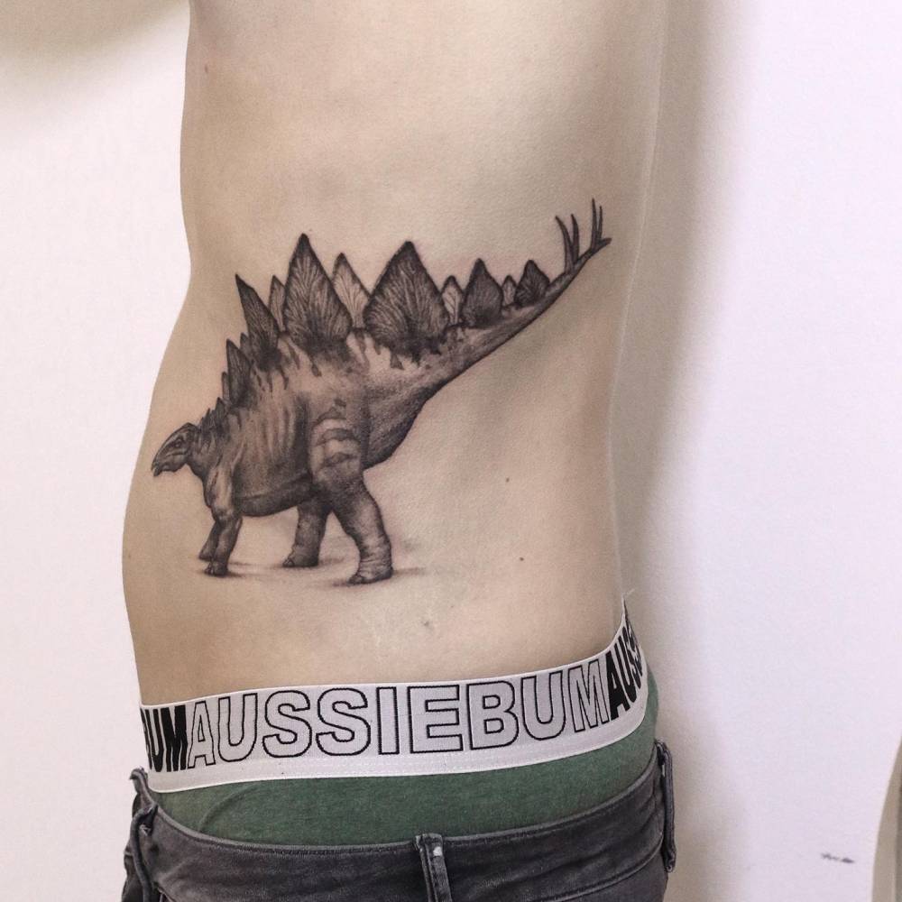 Black and grey stegosaurus tattoo located on the waist.