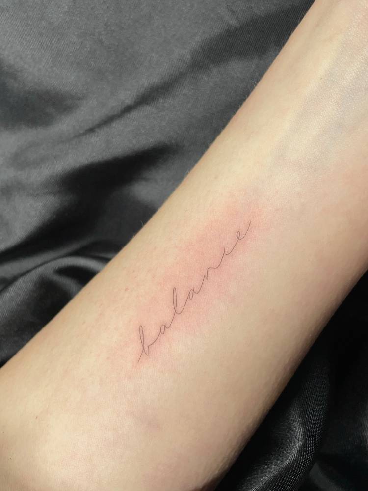 Handwritten font word "balance" tattooed on the inner forearm.