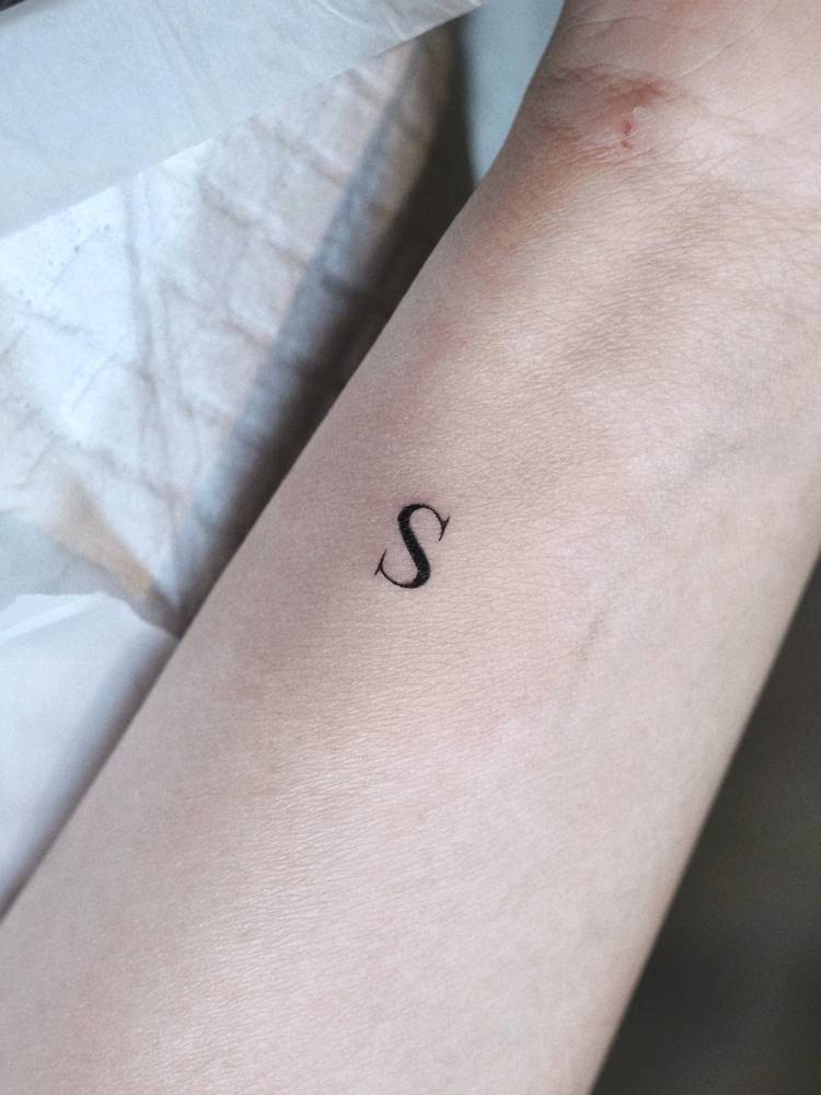 Tiny letter "S" in serif font tattooed on the wrist.