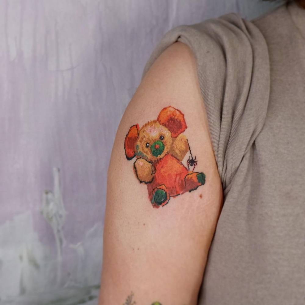 Watercolor style teddy bear tattoo located on the upper arm.