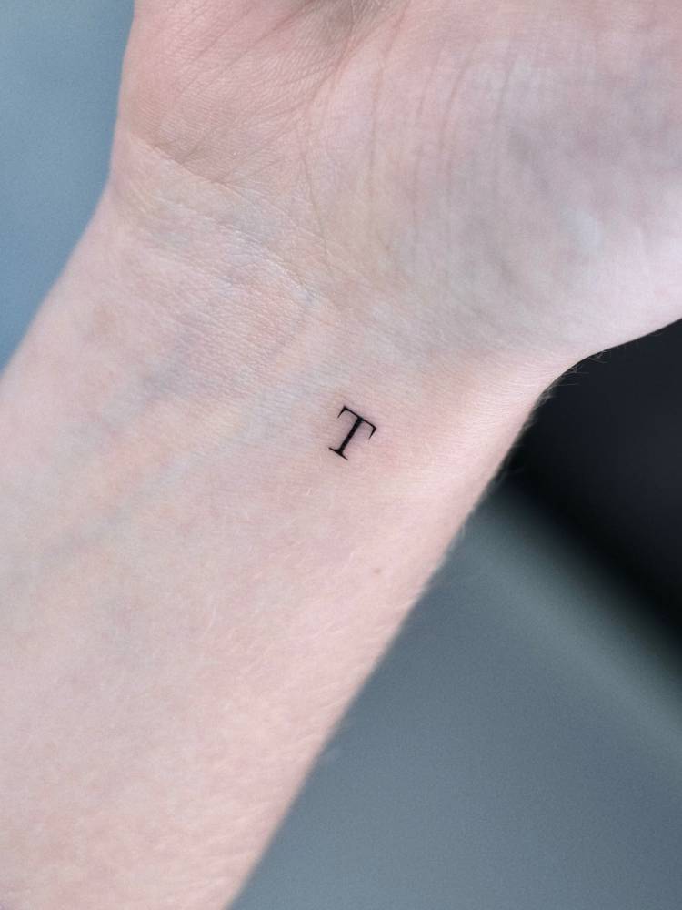 Tiny letter "T" in serif font tattooed on the wrist.