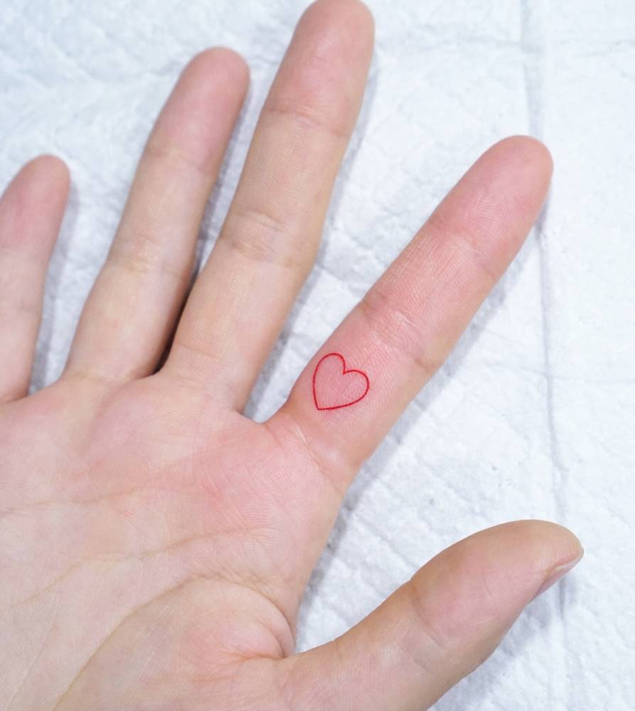 Tiny red heart tattooed on the finger, done in minimalistic style.