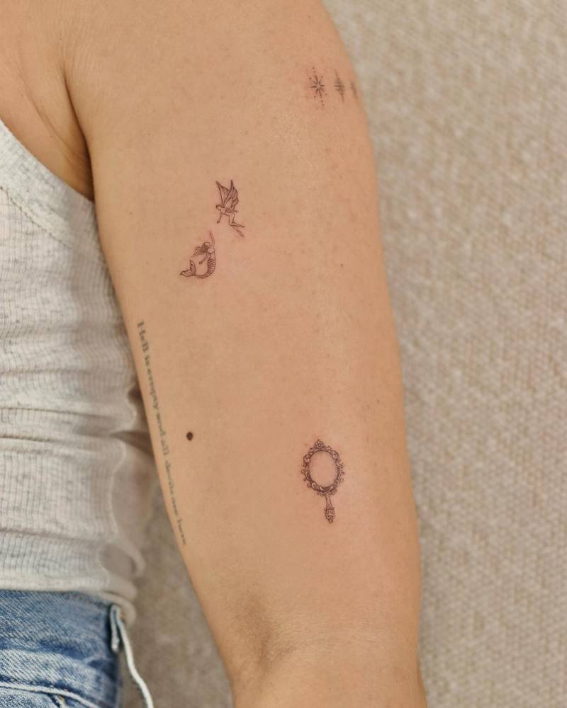 Fine line style mermaid, fairy and mirror tattoos located on the upper arm.