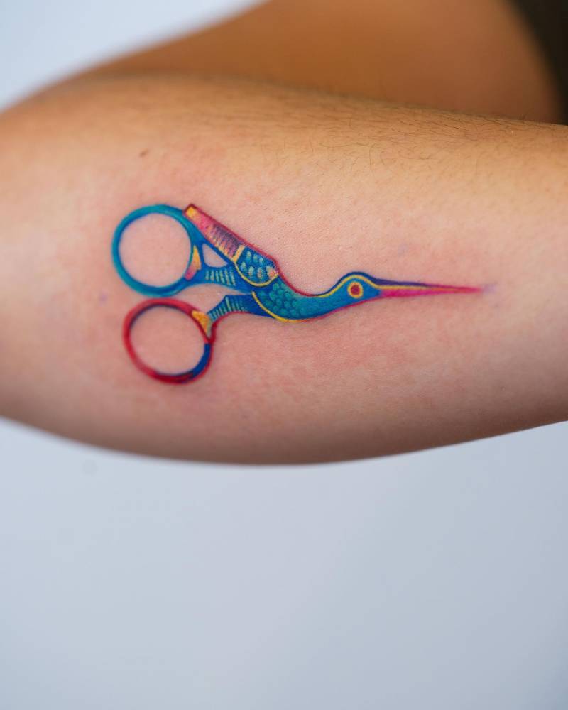 Stork scissors tattoo placed on the forearm, watercolor style.