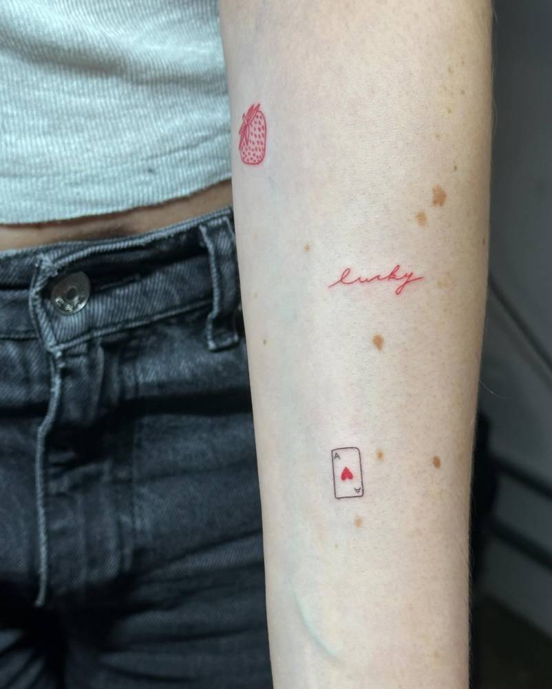 Word "lucky" in red ink and ace of hearts tattoos located on the inner forearm.