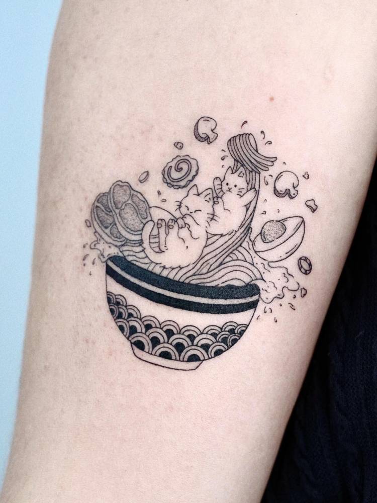 Cartoon style cat in ramen tattoo located on the upper arm.