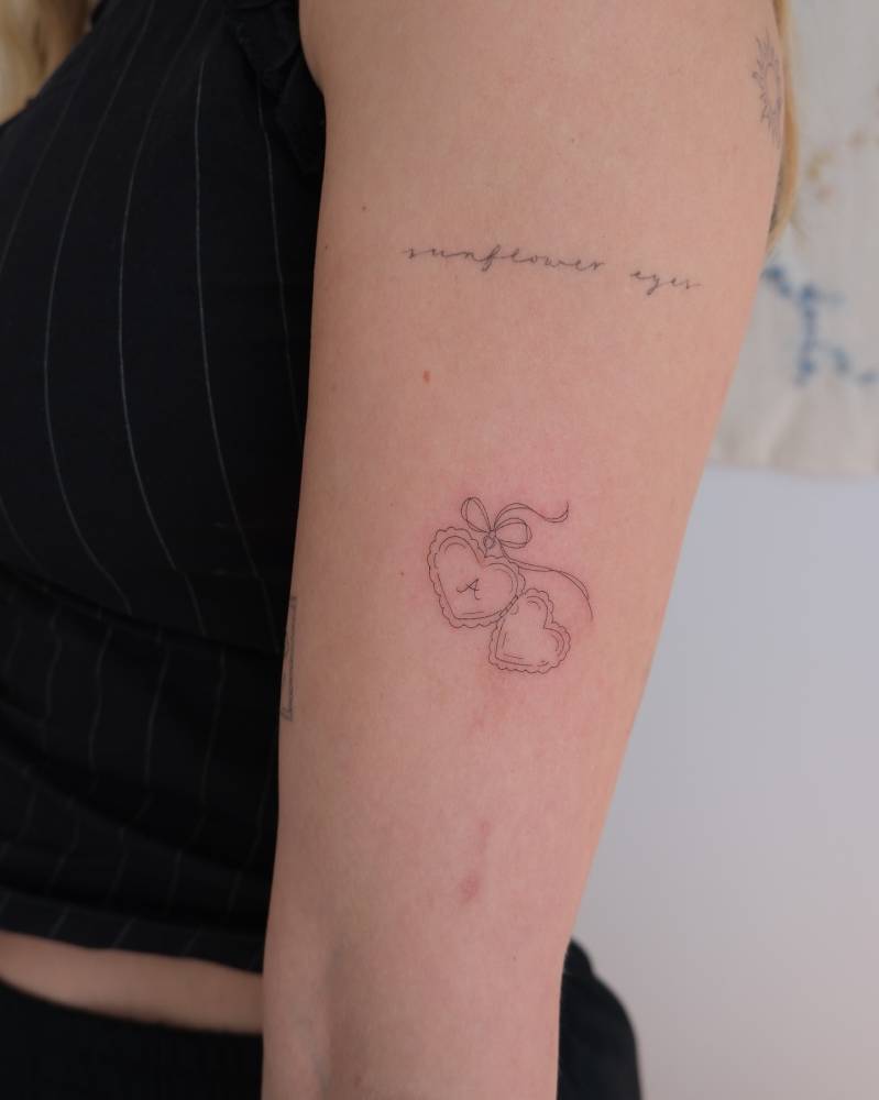 Heart pendant with the letter "A" tattooed on the upper arm, fine line style.