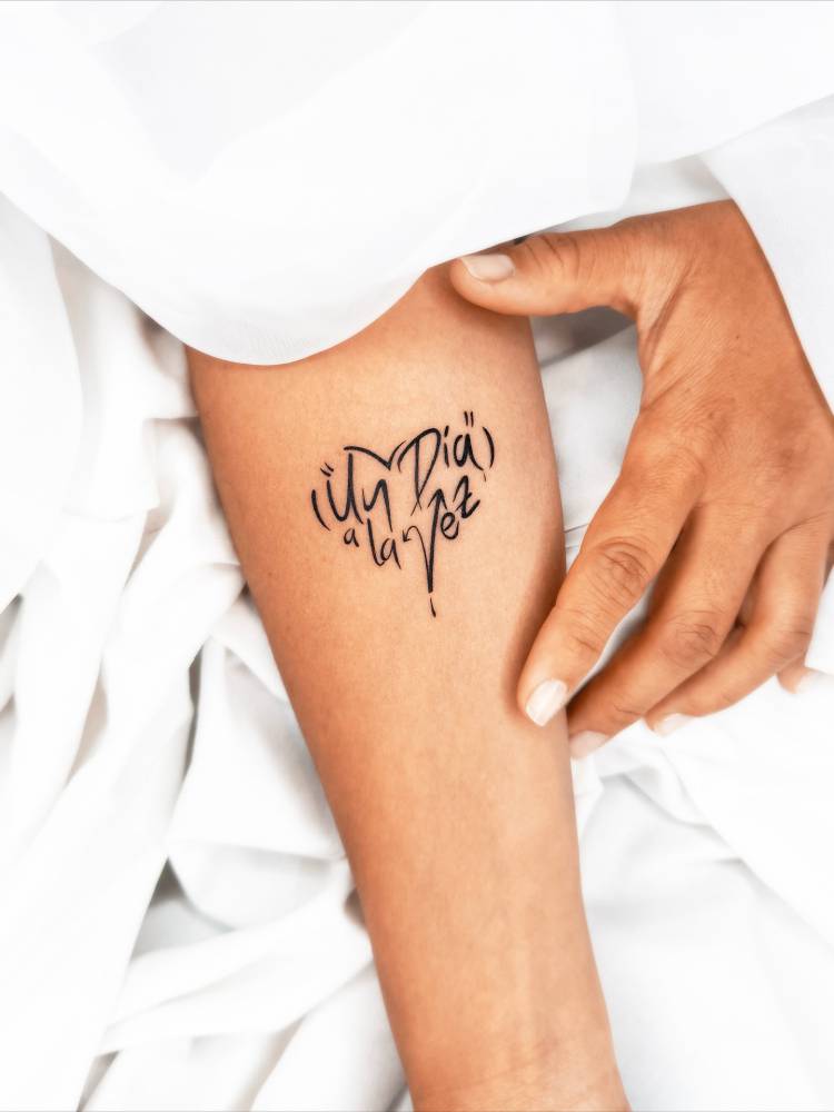 "Un día a la vez" lettering tattoo in a heart shape located on the inner forearm.